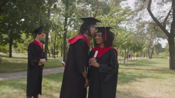 Handsome Arab Male Graduate Whispering to Lovely Black Female Student Outdoors