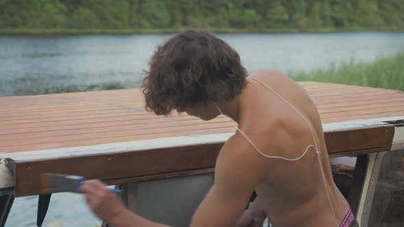 Young topless man painting sliding door overhang on wooden boat to weather-seal.