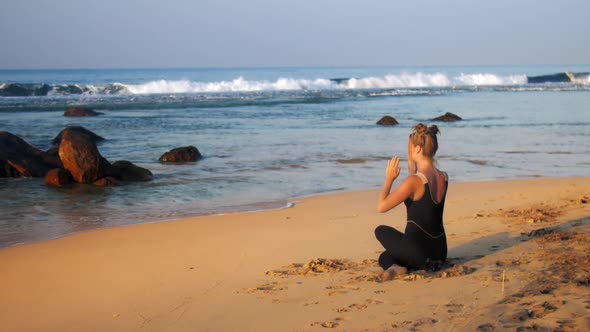 Young Blond Woman in Black Tracksuit Meditates in Yoga Pose