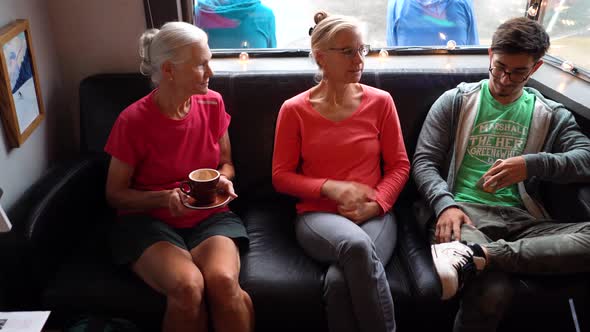 Two mature women talk intimately on a couch as teen son holding smartphone smirks.