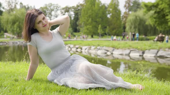 A Young Caucasian Woman Enjoys the Sun with a Smile As She Sits in Grass By a Pond in a City Park