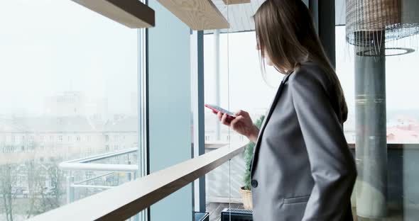 Portrait of Young Attractive Woman Looking at Smartphone on the Office Near Bif Window