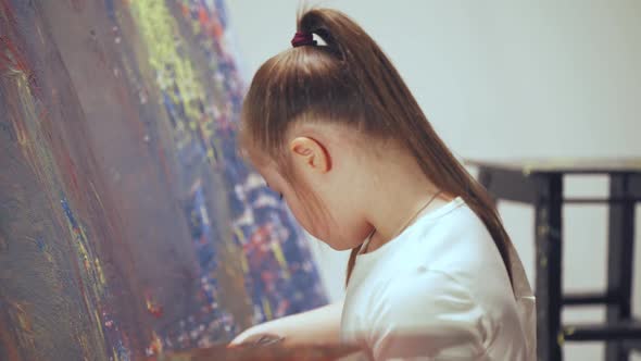 Kid Girl with Down Syndrome Draws with a Brush on a Large Canvas in a White Room Girl with Special