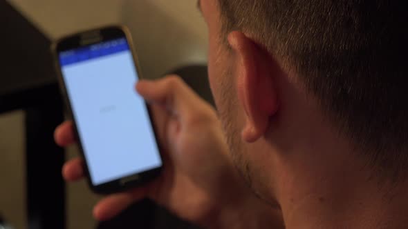 A Man, Turned with the Back of His Head To the Camera, Looks at Pictures of a Woman on a Smartphone