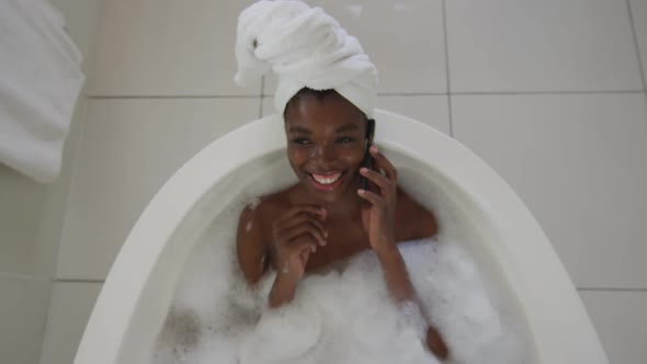 Smiling african american attractive woman relaxing in bath and talking by smartphone in bathroom