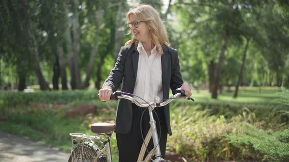 Portrait of Smiling Blond Middle Aged Woman in Suit Sitting on Bike and Leaving