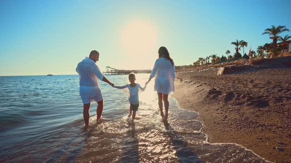 Back View Happy Family Holding Hands and Together Walks Barefoot Along Seashore in Water Waves with