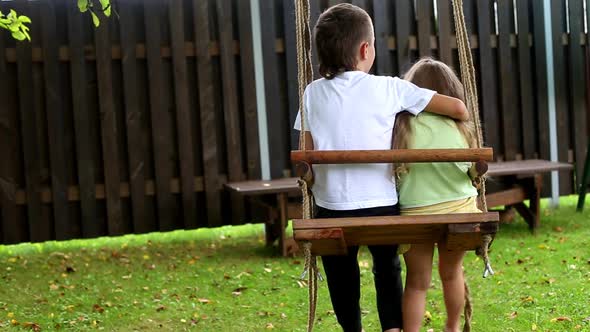 boy and girl ride on a swing in the village. older brother hugging little sister.