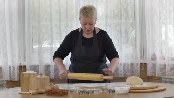 Middle-aged Woman Rolls Dough with Wooden Rolling Pin on Flour Sprinkled Table