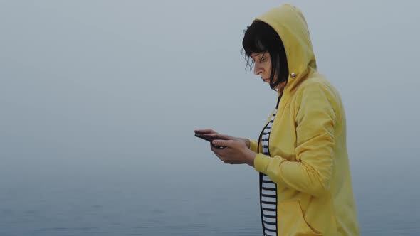 Sleepy Woman Uses Smartphone for Navigation in Foggy Morning By the Water