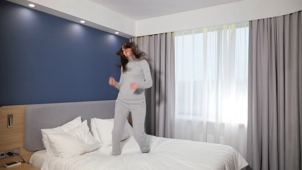 Joyful Girl Jumps on Large Bed Against Window in Hotel Room