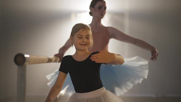 Smiling Happy Girl in Tutu Rehearsing Third Ballet Position with Blurred Woman at Background