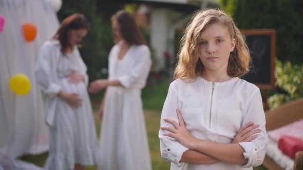 Sad Teenage Girl with Crossed Hands Looking at Camera Sighing As Pregnant Mother Talking to Friend