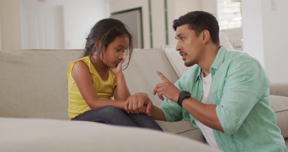 Hispanic father telling off his young upset daughter sitting on sofa