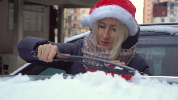 young beautiful Caucasian woman in Santa hat cleans car from snow with a brush