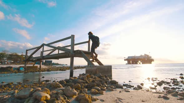 Surfer guy walks up wooden bridge with oil platform in background, Curacao