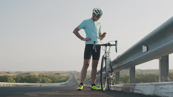 A guy in tight shorts, a helmet, and sunglasses is standing on the side of the road near the bike an