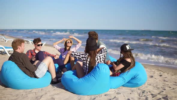 Young Man Playing Guitar Among Group of Friends Sitting on Easychairs on the Beach and Singing on a