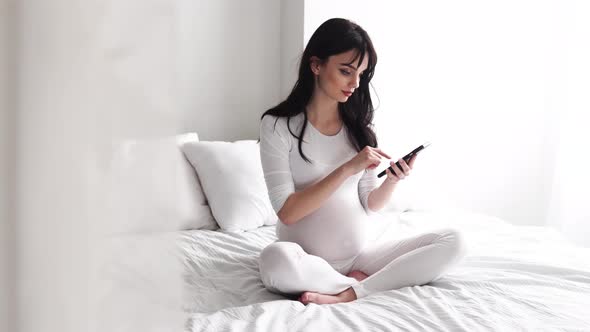 Pregnant Woman With Mobile Phone Sitting On Bed At Bedroom