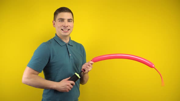 A Young Man Inflates a Long Red Balloon with a Pump on a Yellow Background. Man in a Green Polo.