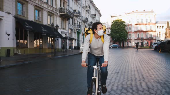 Courier with Yellow Backpack and Protective Mask Rides a Bicycle Through the City with Food Delivery