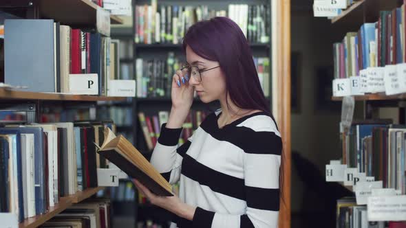 Female student with dark hair adjusts her glasses and reads a book, standing in the library, between