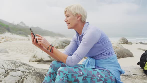 Senior hiker woman with backpack using smartphone while sitting on rock hiking on the beach.