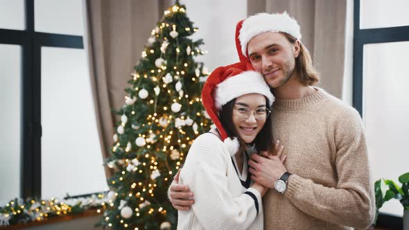 Multiracial Pair in Santa Hats are Smiling and Hugging Each Other Standing at Home Against Green