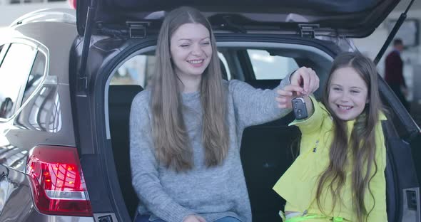 Young Caucasian Woman and Little Cute Girl Sitting in Car Trunk and Bragging Keys. Positive Mother