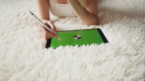 Woman illustrator lies on carpet and draws with stylus on graphic tablet with chromakey screen