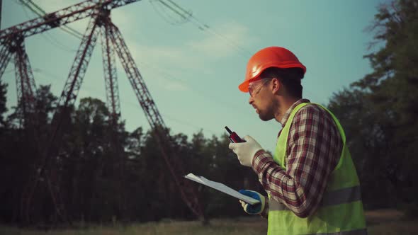 Caucasian Engineer in Protective Uniform and Orange Hard Hat Holds Walkietalkie and Clipboard on