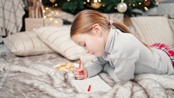 Cute Little Girl Writing Letter to Santa Claus Lying on Floor Near Christmas Tree at Home Tracking