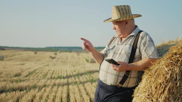 Old Farmer Uses Tablet in the Field Next to Haystack at Sunset