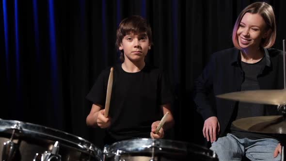 Young Caucasian Woman Teaches a Boy to Play the Drums in the Studio on a Black Background