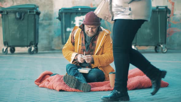 A Poor Man is Eating While Sitting Next to the Trash Bins