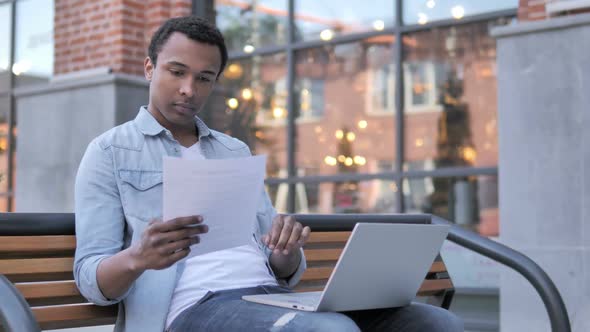 African Man Reading Contract and Working on Laptop Sitting on Bench