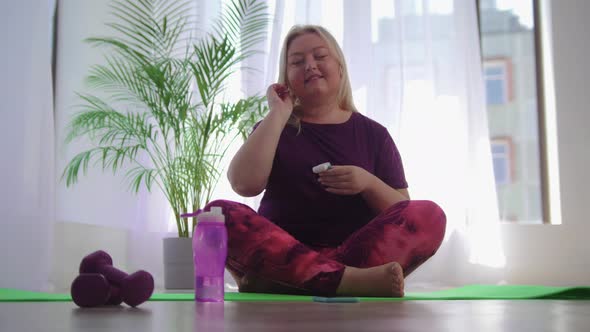 Fitness Training  Blonde Overweight Woman Sitting on Yoga Mat and Puts on Her Wireless Headphones