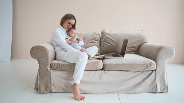 Pleasant Young Mother Freelancer Working on Computer Sitting on a Sofa with Cute Baby Infant at Home