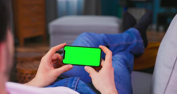 Young Man at Home Resting on a Couch Using with Green Mockup Screen Cell Phone