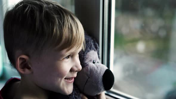 Little Boy Smiling Looking Out the Window Child Looks Out the Window on a Rainy Day alt