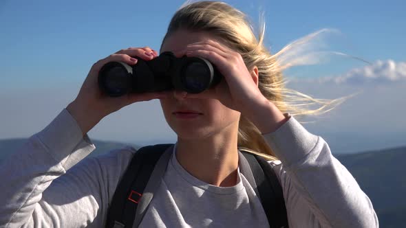 A Young Woman on a Hilltop Looks Around Through Binoculars - Closeup - Mountainous Landscape