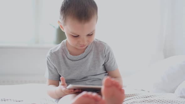 A Small Caucasian Boy in a Light Room in a Gray Tshirt Plays with a Tablet While Sitting on the