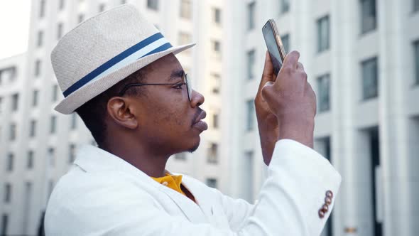 Concentrated black guy in classic suit films highrise office buildings holding smartphone