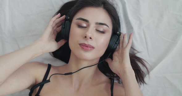 Woman Lying on Bed Holds Earcups Sways to Music Looks with Smile at Camera