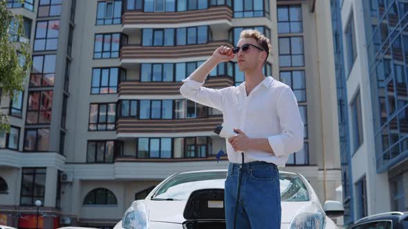 Stylish Modern Young Curly Man in Jeans and a White Shirt Stands Near an Electric Car with a Charger