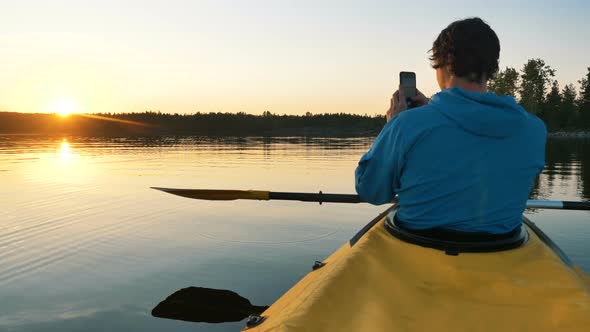 Man Takes a Photo of Sunset on Smartphone on Lake on Kayak with Paddle, Self-isolation Outdoor