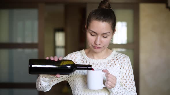 girl pours red wine into a mug and drinks, looking out the window in the kitchen