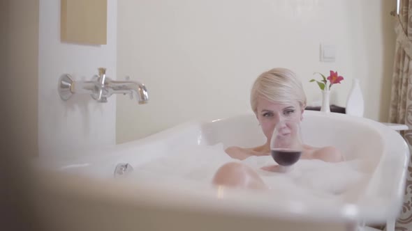 Relaxed Blond Woman Drinking Red Wine in Bathtub with Bath Foam. Portrait of Happy Caucasian Lady