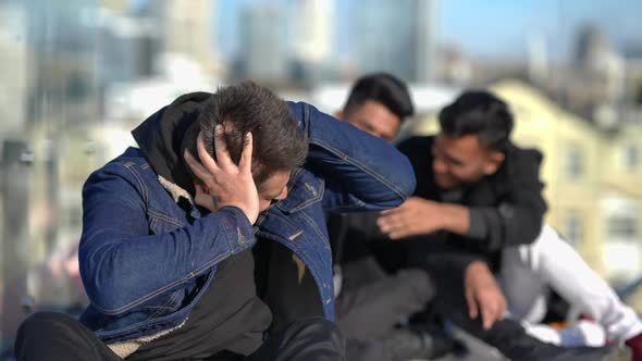 Front View Depressed Bullied Young Man Holding Head in Hands As Blurred Mates Pointing Laughing