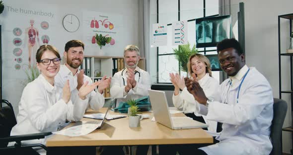 Multicultural Male and Female Doctors Sitting Around Workplace in Clinic and Clapping Hands